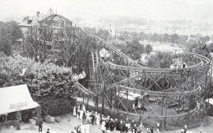 A historic rollercoaster surrounded by residential buildings and green countryside, with a queue of guests.