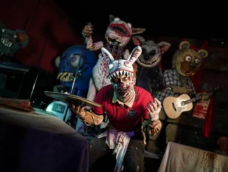 Traumatica Charakter Zombie-Kellner und fünf Plüschmonster Traumatica Charakter Zombie-Kellner mit gruseliger Hasenkopf-Mütze hält ein Tablett in seiner Hand. Im Hintergrund sind riesige Plüschmonster mit großen Fangzähnen.