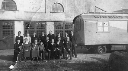 The Mack family and employees stand in front of the wagon factory building, with circus wagons in the background.