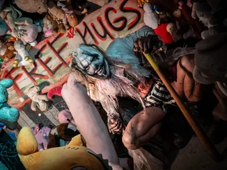 Traumatica Clown bietet "Free Hugs" Traumatica Charakter mit hässlicher Clownsfratze steht vor einer Wand voller Puppen und Plüschtiere. In der Mitte steht ein Schild mit der Aufschrift Free Hugs.