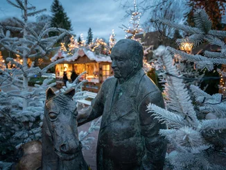 Statue of Franz Mack in a winter setting with lights and a Christmas market at dusk. Bronze statue of Franz Mack with a horse between decorated Christmas trees at the Europa-Park Christmas market.