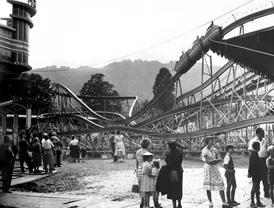 A group of people stand in front of a large rollercoaster in a field, against the backdrop of a mountain landscape.