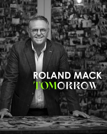 Schwarz-weiß Aufnahme von Roland Mack vor einer Wand voller Fotos.