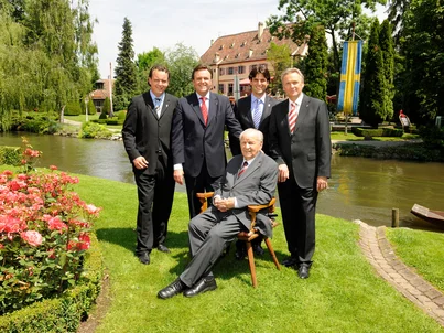 Franz Mack is sitting on a lawn in Europa-Park. Roland, Michael, Thomas and Jürgen Mack are standing behind him, with Balthasar Castle in the background.