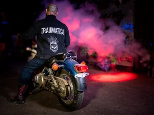 Model mit Traumatica Jeansjacke Mann trägt Traumatica Jeansjacke mit Aufschrift, sitzt auf Motorrad.